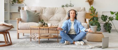 Kulaklıklı genç bir kadın oturma odasında meditasyon yapıyor.