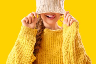 Sarı arka planda dilini gösteren kalın süveter ve şapkalı güzel, mutlu genç bir kadın.