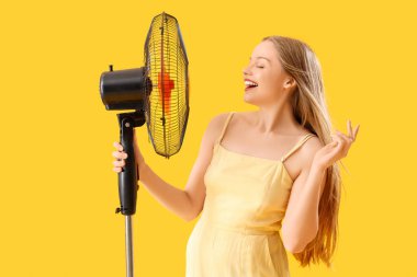 Young woman with electric fan on yellow background clipart