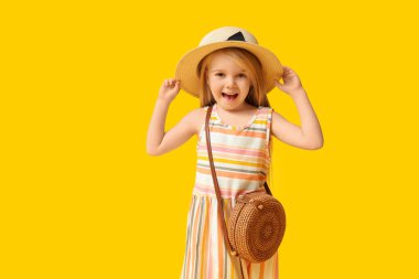 Cute little girl in hat with stylish wicker bag on yellow background