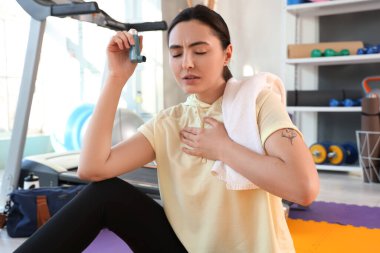 Sporty young woman with inhaler and towel having asthma attack in gym clipart