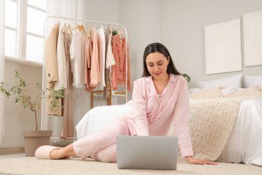 Yatak odasındaki elbise rafının yanında dizüstü bilgisayar kullanan genç bir kadın.
