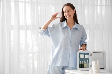 Evinde küçük kozmetik buzdolabı ve krema dolu bir kavanozla masanın yanında oturan mutlu genç kadın.