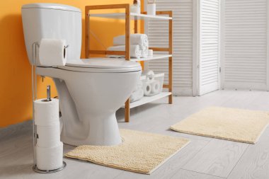 Tuvaletin içinde klozet, raf ünitesi ve sarı duvarın yanında kağıt tutucu var.