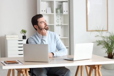 Bilgisayarları olan düşünceli genç işadamı masadaki masaya doğru bakıyor.
