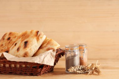 Lezzetli lavaş parçaları, buğday dikenleri ve hafif ahşap arka planda baharat kavanozlarıyla dolu bir sepet.