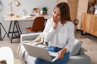 Dizüstü bilgisayarlı genç bir kadın evdeki kanepede sohbet ediyor.