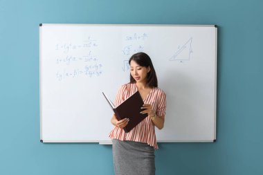 Sınıfta yazı tahtasının yanında kitabı olan Asyalı Matematik öğretmeni