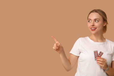 Young woman with blisters of vitamin A pills pointing at something on beige background clipart