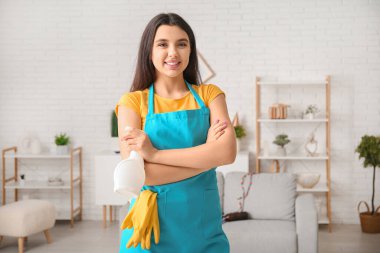 Oturma odasında elinde sprey deterjan şişesi olan genç bir kadın.