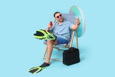 Şişme yüzüklü, kokteyl ve evrak çantalı mutlu bir ofis çalışanı şezlongda oturuyor ve mavi arka planda cep telefonuyla selfie çekiyor.