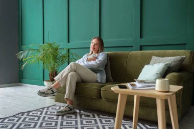 Genç bir kadın yeşil oturma odasında yastıkla kanepede oturuyor.