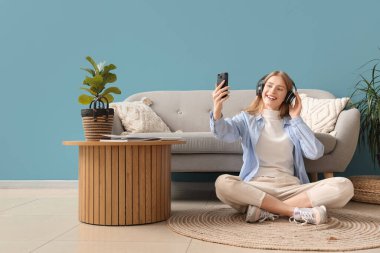Kulaklıklı genç ve güzel bir kadın mavi oturma odasında cep telefonuyla selfie çekiyor.