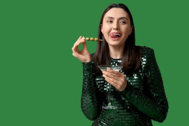 Güzel genç bir kadın elinde zeytinli bir bardak martini tutuyor ve yeşil arka planda dilini gösteriyor.