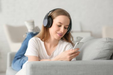 Kulaklıklı, müzik dinleyen ve oturma odasındaki kanepede uzanan genç ve güzel bir kadın.