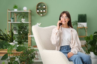 Evdeki çiçeklerin yanındaki koltukta cep telefonuyla konuşan genç bir kadın.