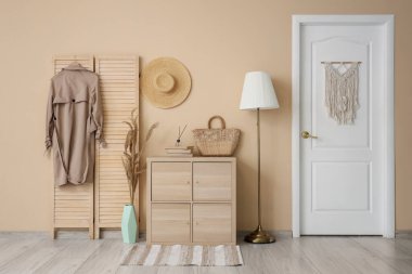 Katlanan ekrandaki trençkot, koridordaki bej duvarın yanında çanta ve lambayla dolu çekmeceler.