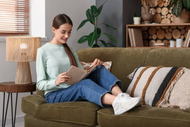 Oturma odasında kitapla kanepede oturan genç kadın.