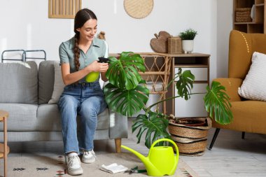 Genç bir kadın oturma odasındaki Monstera bitkisine su püskürtüyor.