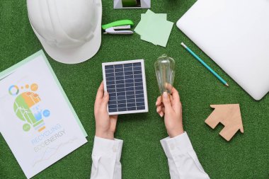 Ampulü, güneş paneli, ev figürü ve çimlerin üzerinde kırtasiye malzemesi olan bir kadın. Yeşil Enerji kavramı