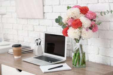 Modern dizüstü bilgisayar, bir fincan kahve, güzel yıldız çiçekleri ve ışıklı tuğla duvarın yanındaki masada kırtasiye malzemeleri.