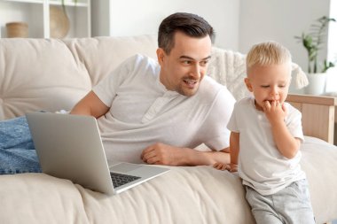 Baba kanepede yatar ve dizüstü bilgisayarında çocuğuna bir şey gösterir.