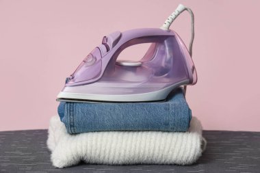 Odasında elektrikli ütü ve pembe duvarın yanında giysi yığını var.