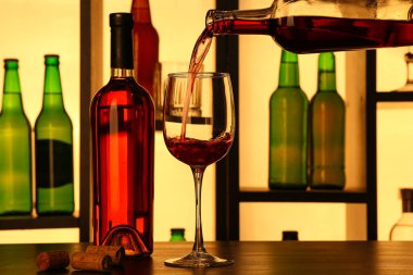 Barda şişeler olan rafların yanındaki bardaklara kırmızı şarap dökülüyor.