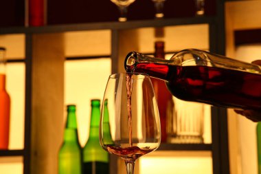 Barda şişeler olan rafların yanındaki bardaklara kırmızı şarap dökülüyor.