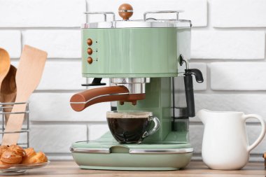 Modern kahve makinesi, bir bardak sıcak espresso ve bir tabak tatlı keki, mutfaktaki beyaz tuğla duvarın yanındaki ahşap masada.