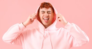 Pembe arka planda dil gösteren kapüşonlu komik kızıl saçlı genç adam.
