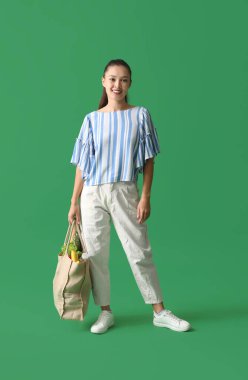 Yeşil arka planda bir çanta dolusu taze yiyecek taşıyan genç Asyalı kadın.