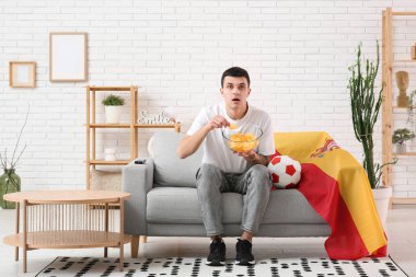 Bir kase patates cipsi, futbol topu ve İspanya bayrağıyla evdeki kanepede futbol izleyen genç adam.