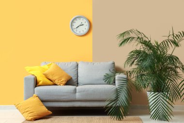 Şık bir oturma odası, rahat bir kanepesi, sarı duvarda çiçekleri ve saati var.