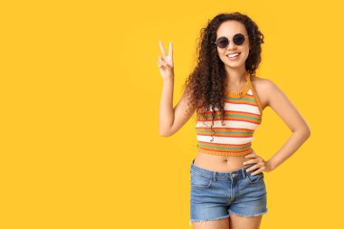 Güneş gözlüklü güzel Afro-Amerikalı kadın sarı arka planda zafer işareti gösteriyor.