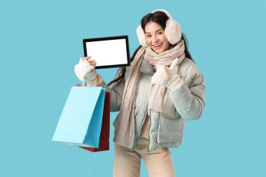 Mavi arka plandaki modern tablet bilgisayarını işaret eden alışveriş torbalarıyla kalın giysiler içinde güzel bir genç kadın.