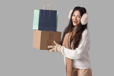 Gri arka planda, alışveriş torbaları ve karton kutusuyla kalın giysiler içinde güzel bir kadın.