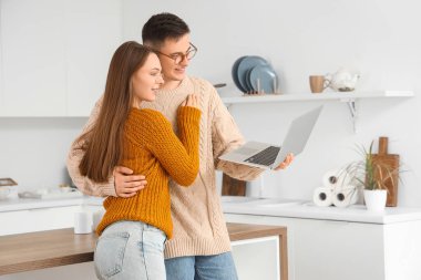 Evde dizüstü bilgisayarlı ve sıcak kazaklı sevimli genç çift sarılıyor.