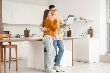 Evde dizüstü bilgisayarlı ve sıcak kazaklı sevimli genç çift sarılıyor.