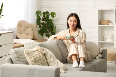 Oturma odasındaki kanepede bir fincan sıcak çayla oturan güzel, genç Asyalı kadın.