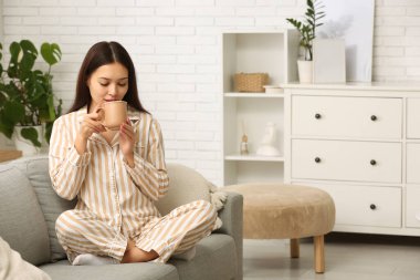 Güzel genç Asyalı kadın sıcak çay içiyor ve oturma odasında kanepede oturuyor.