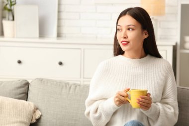 Oturma odasındaki kanepede bir fincan sıcak çayla oturan güzel, genç Asyalı kadın.