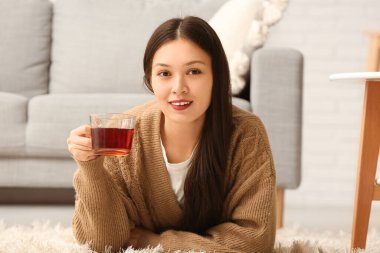 Güzel genç Asyalı kadın, oturma odasında sıcak çay bardağıyla halıya uzanmış.