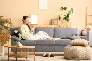 Oturma odasındaki kanepede bir fincan sıcak çayla oturan güzel, genç Asyalı kadın.