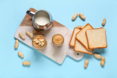 Fıstık ezmeli kavanozlar, bir sürahi süt, kızarmış ekmek ve fındıklar mavi arka planda.