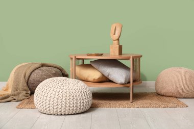 Yeşil duvarın yanında yastıklar ve puflar olan kahve masası.