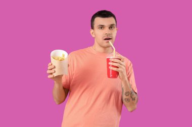 Genç adam kola içiyor ve mor arka planda patates kızartması yiyor.