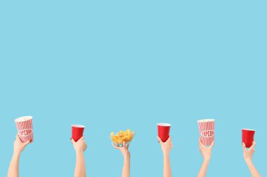 Mavi arka planda patlamış mısır, içecek ve patates cipsi dolu kovaları tutan eller.