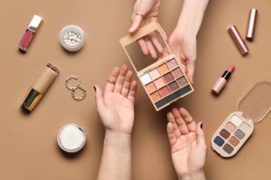 Farklı dekoratif kozmetik ürünleri ve kahverengi arka planda küpeleri olan kadın elleri.