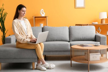 Genç kadın dizüstü bilgisayar kullanıyor ve güzel oturma odasındaki kanepede oturuyor.